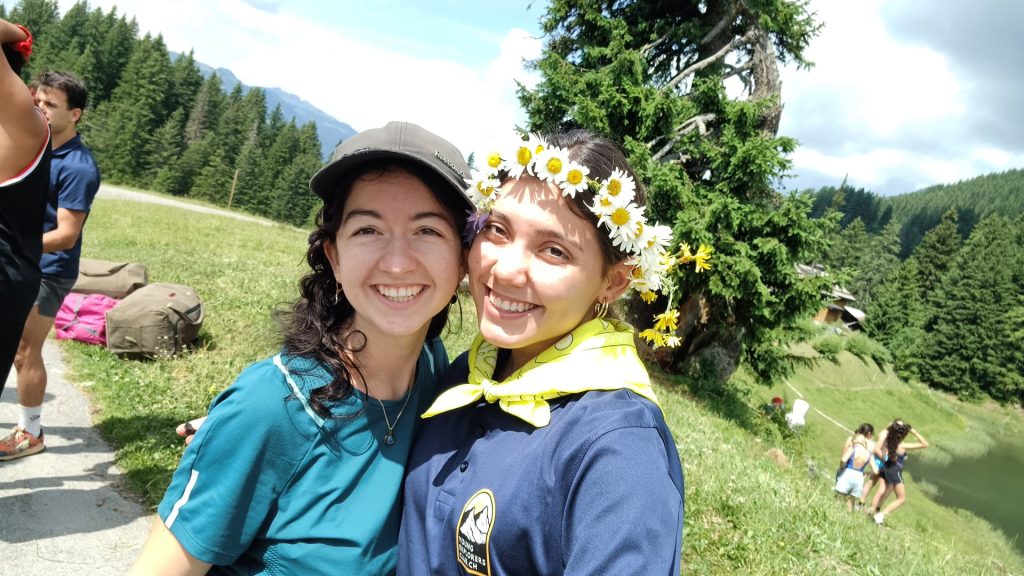 Deux monitrices souriantes, l'une avec une couronne de marguerites, au bord d'un lac alpin