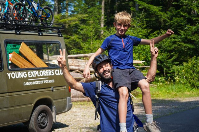 Moniteur portant un jeune campeur sur le dos à côté du van Young Explorers Club chargé de vélos