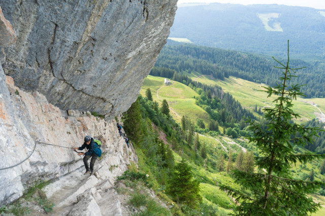 Climber traversing a via ferrata cable along an overhanging cliff above the valley