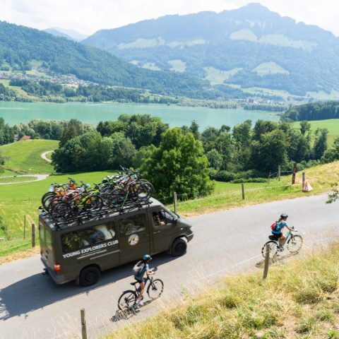 Young Explorers Club support van loaded with bikes escorting young cyclists along a lakeside road