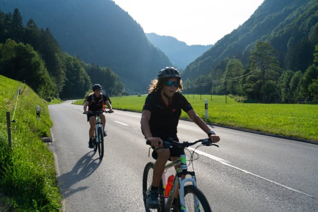 Two young cyclists riding along a valley road between forested mountains