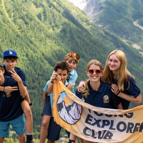 Teenagers holding a Young Explorers Club banner on a mountain pass during the Tour du Mont Blanc camp