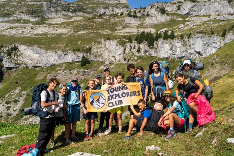 Group of teenagers with hiking backpacks holding the Young Explorers Club banner below limestone cliffs