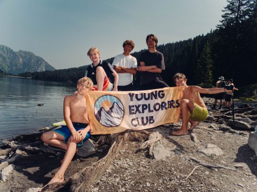 Jugendliche halten das Young Explorers Club Banner an einem felsigen Seeufer in den Bergen