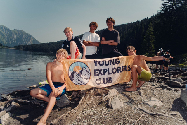 Ados tenant la bannière Young Explorers Club sur la rive rocheuse d'un lac de montagne