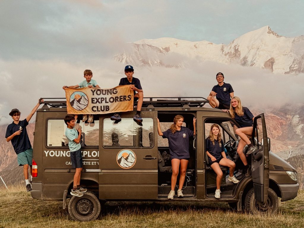 Teenagers and staff on the Young Explorers Club van holding the club banner in the mountains during a travel camp