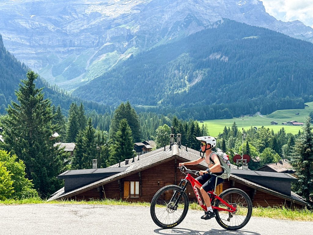 Vététiste passant devant des chalets avec le massif des Diablerets en arrière-plan