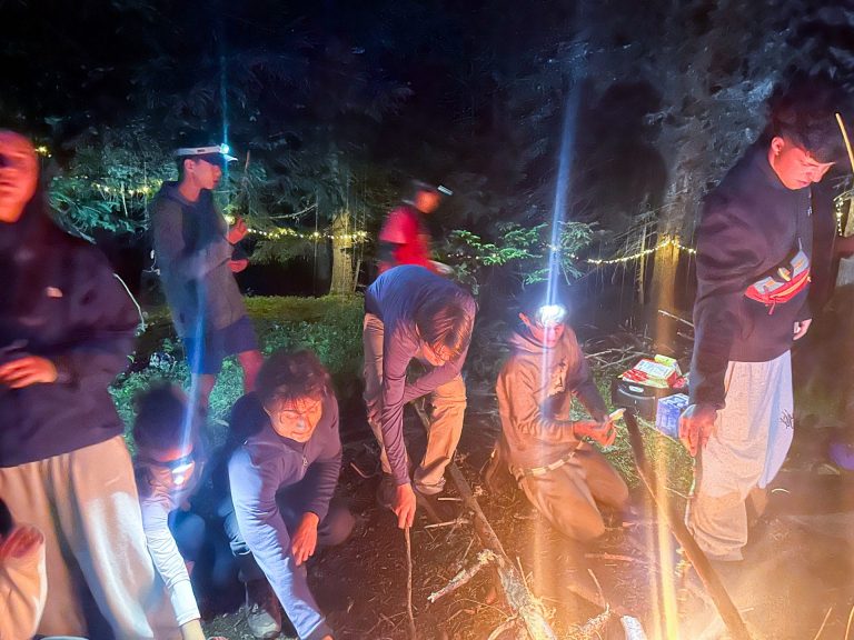 Campistas con frontales reunidos alrededor de una hoguera de noche en el campamento Survival Academy