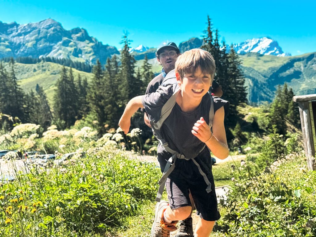 Niño sonriendo mientras baja corriendo un sendero de montaña con cumbres nevadas al fondo