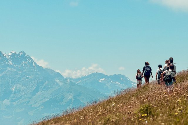Campers hiking up a flower meadow ridge with the snow-capped Alps behind