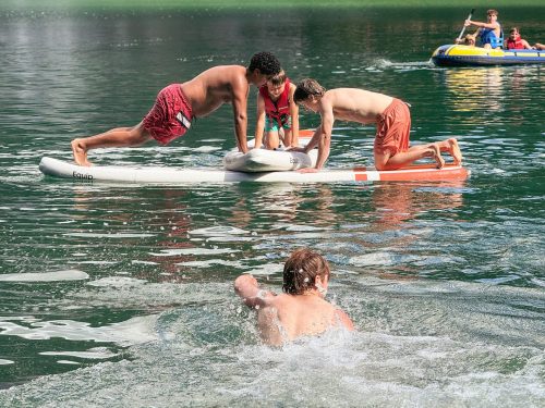 Teenagers balancing on a paddleboard on a mountain lake while a friend swims alongside