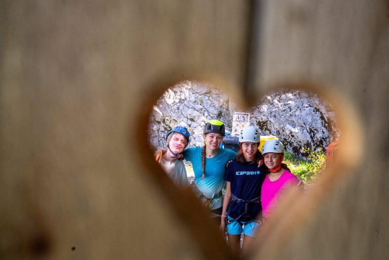 Four campers in climbing helmets seen through a heart-shaped cut-out in a wooden wall