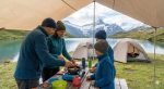 Family preparing a meal under a tarp at a lakeside camp in the mountains